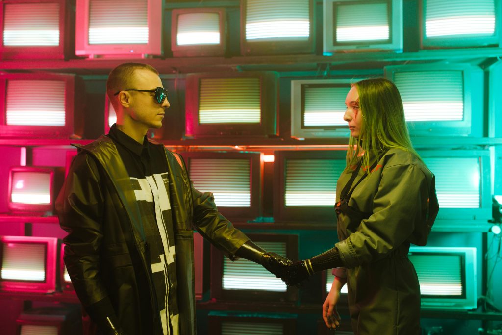 pexels-photo-7688736-7688736 Two individuals with prosthetic arms engage in a handshake amidst a vibrant, retro television backdrop.
